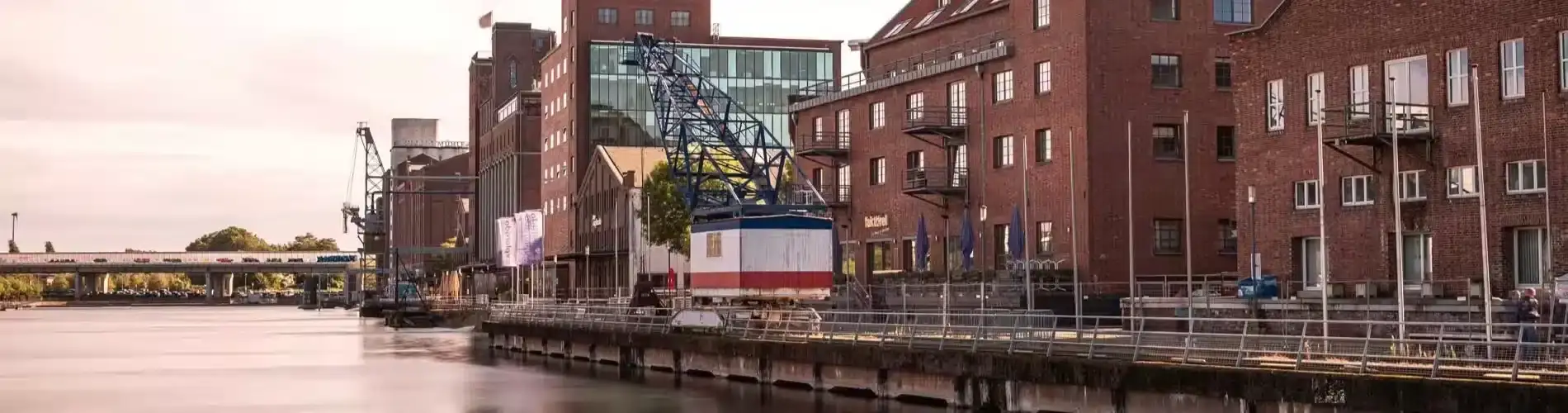 Ein alter Kran steht unbewegt am Ufer eines ruhigen Flusses, umgeben von historischen Backsteingebäuden und einem modernen Gebäude.